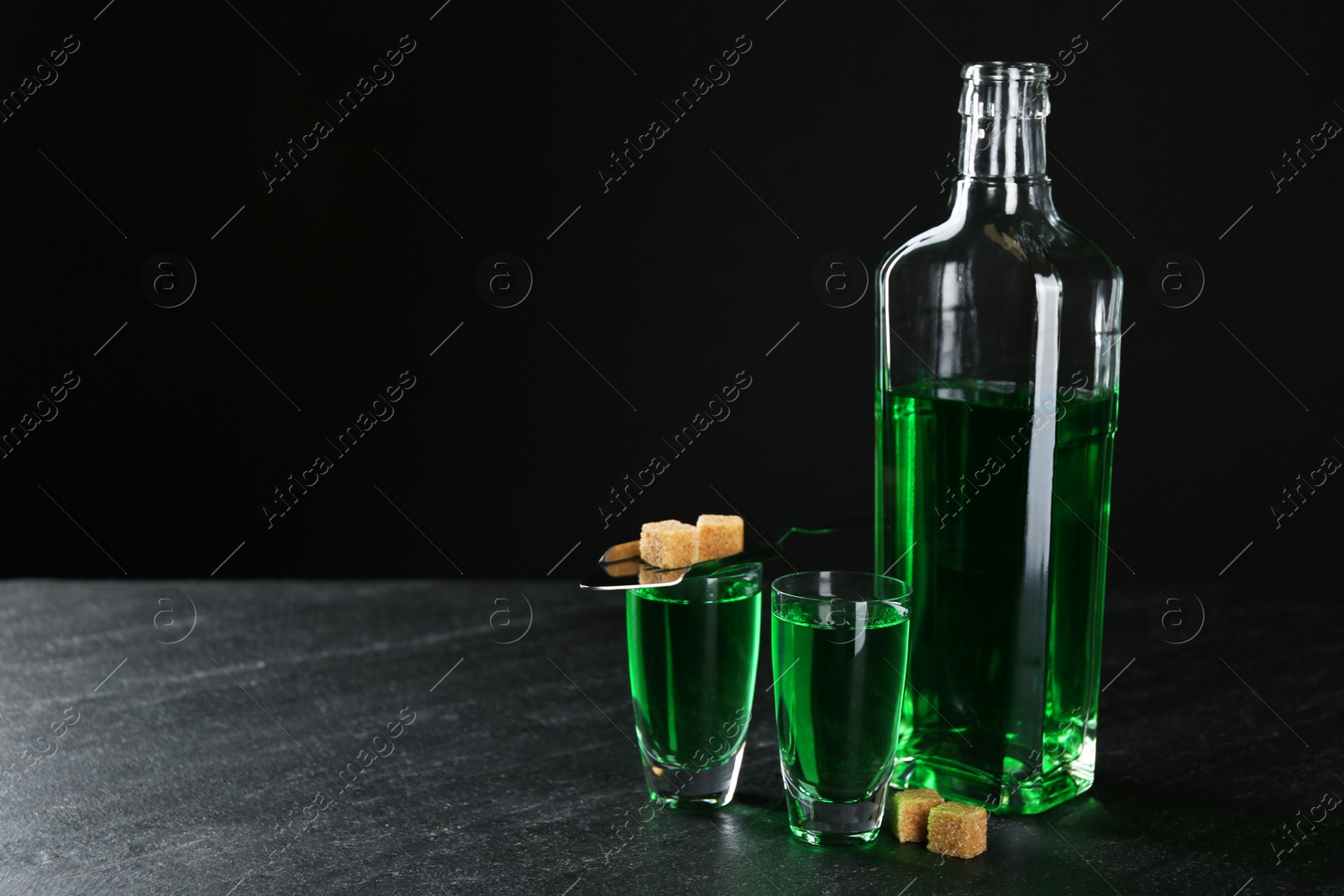 Photo of Absinthe in shot glasses, bottle, brown sugar and slotted spoon on table against black background, space for text