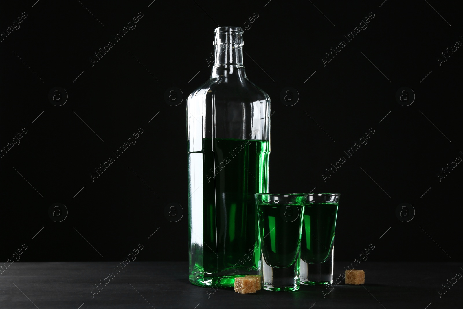 Photo of Absinthe in shot glasses, bottle and brown sugar on table against black background