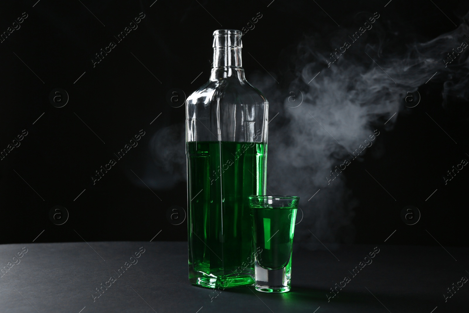 Photo of Absinthe in shot glass and bottle on table against black background with smoke