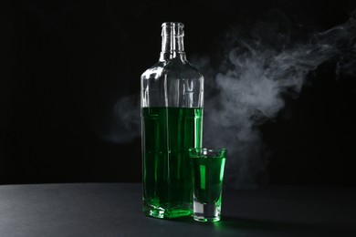 Photo of Absinthe in shot glass and bottle on table against black background with smoke