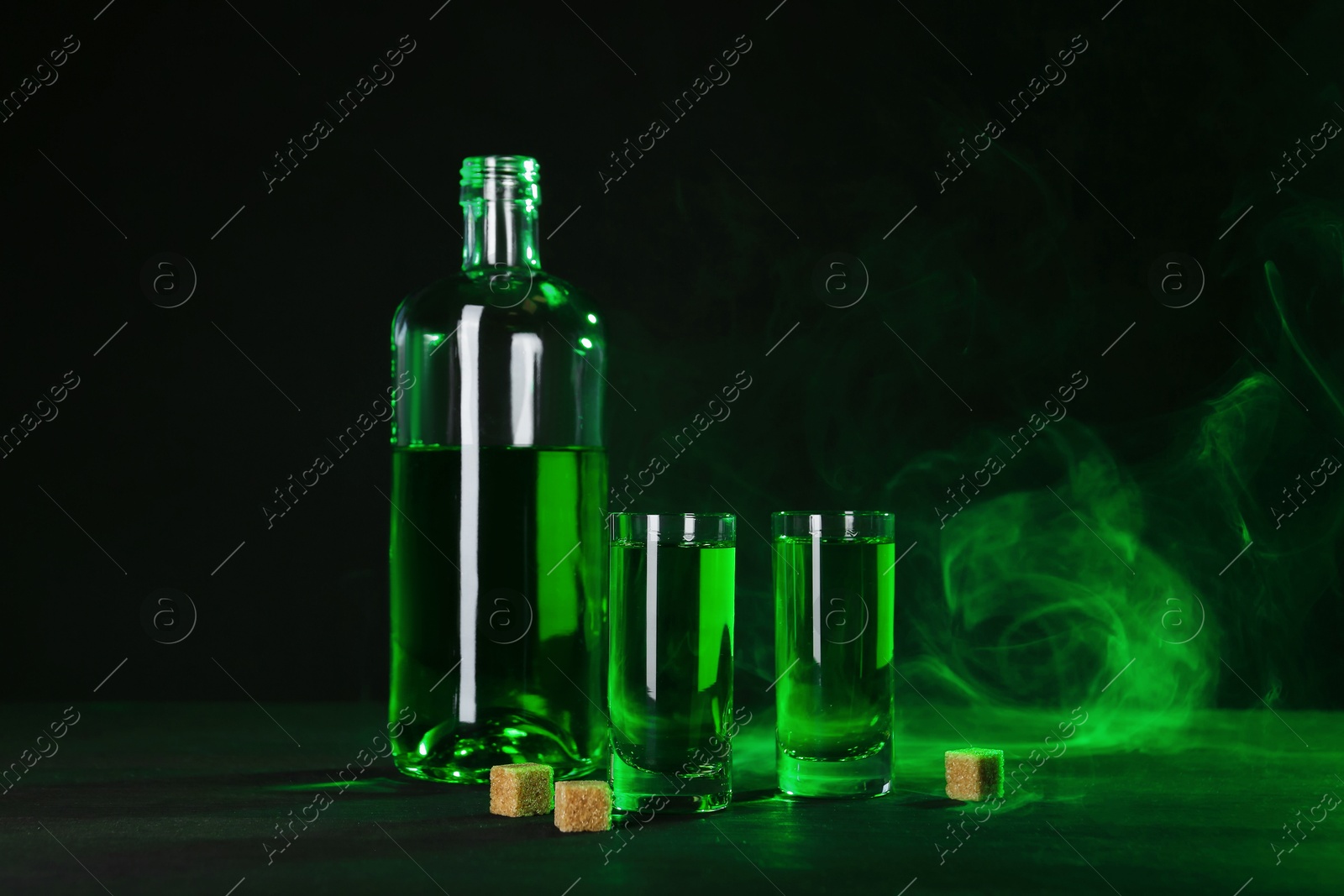 Photo of Absinthe in shot glasses, bottle and brown sugar on table against black background with smoke