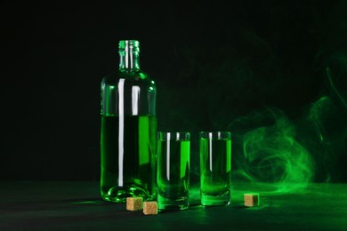 Photo of Absinthe in shot glasses, bottle and brown sugar on table against black background with smoke