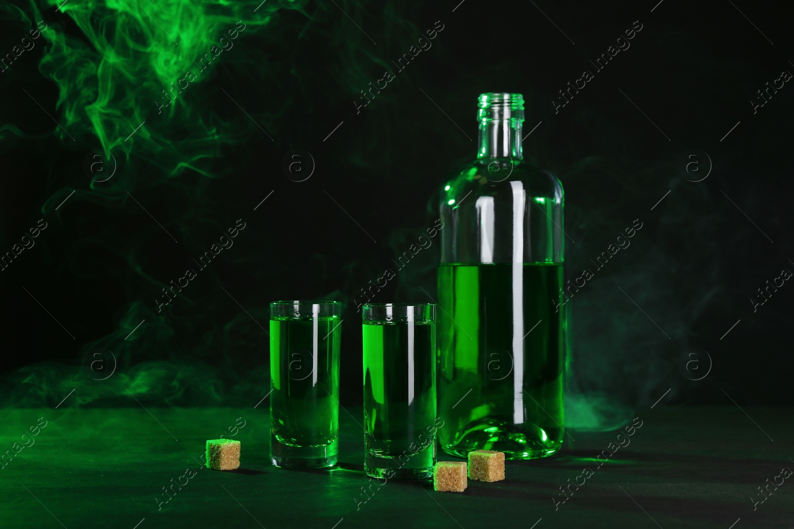 Photo of Absinthe in shot glasses, bottle and brown sugar on table against black background with smoke