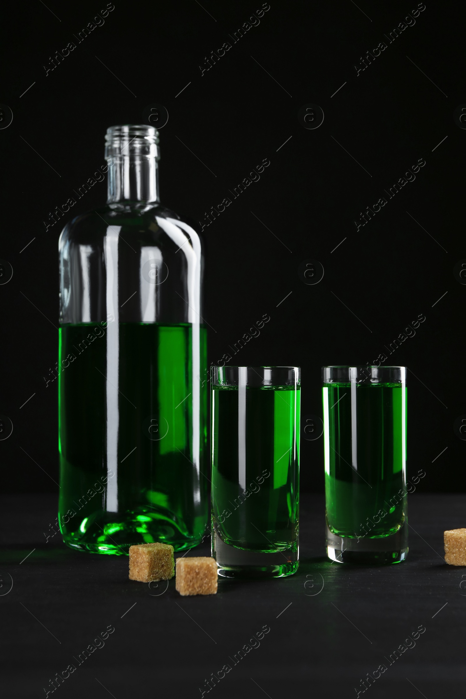 Photo of Absinthe in shot glasses, bottle and brown sugar on table against black background