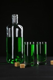 Photo of Absinthe in shot glasses, bottle and brown sugar on table against black background