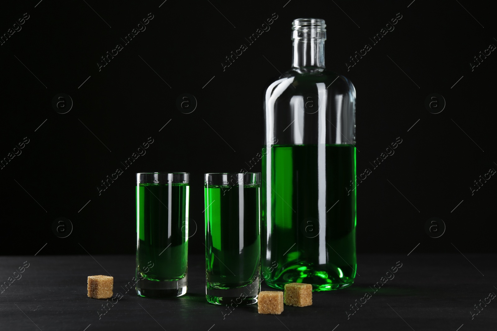 Photo of Absinthe in shot glasses, bottle and brown sugar on table against black background