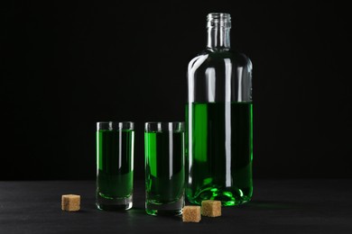 Photo of Absinthe in shot glasses, bottle and brown sugar on table against black background