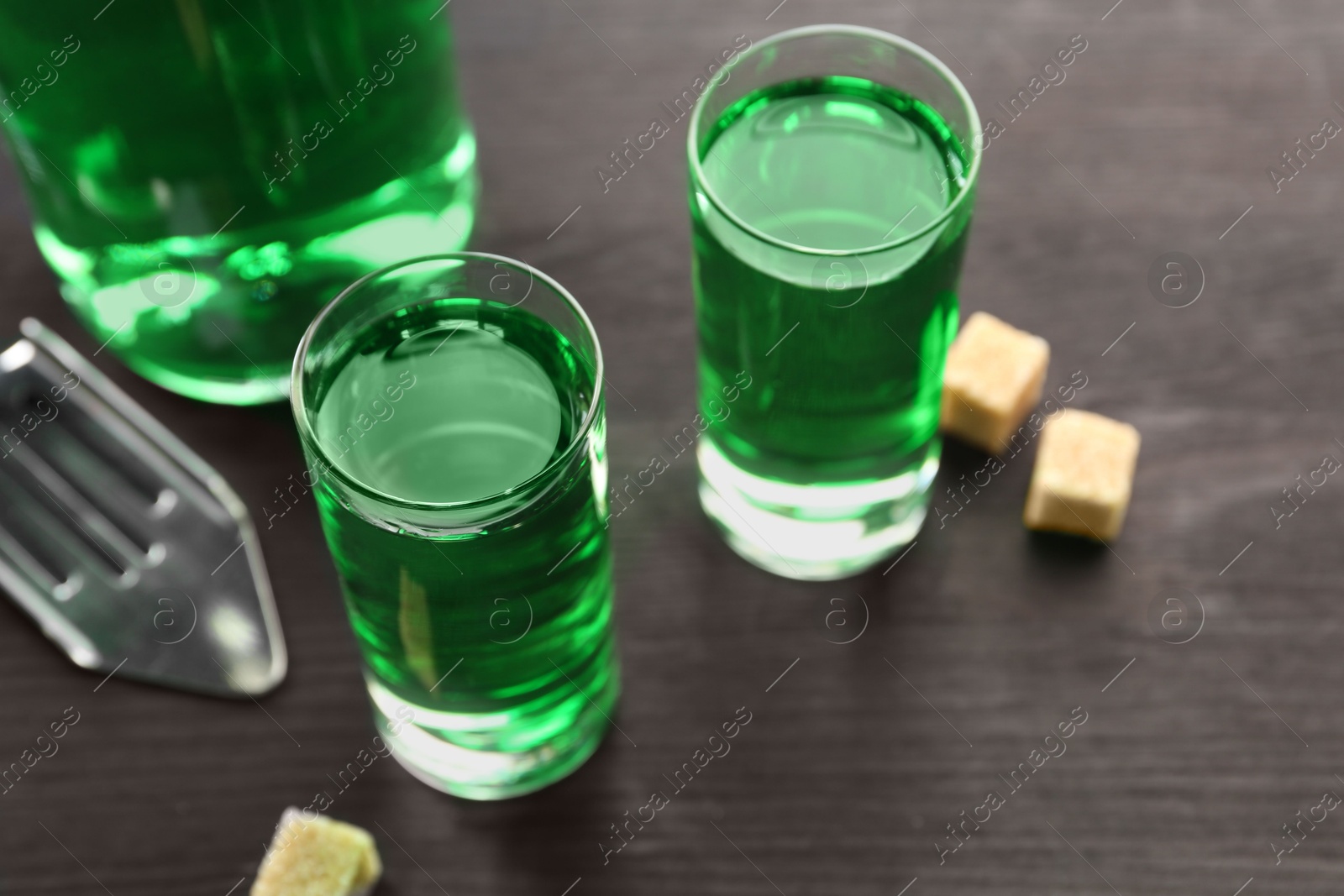 Photo of Absinthe in shot glasses, brown sugar and slotted spoon on wooden table, closeup