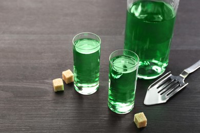 Photo of Absinthe in shot glasses, bottle, brown sugar and slotted spoon on wooden table, closeup. Space for text