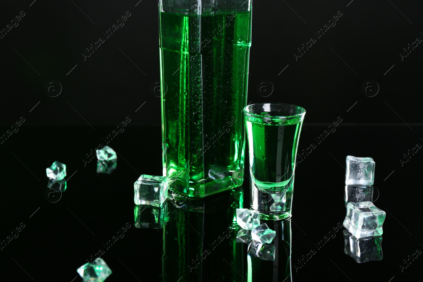 Photo of Absinthe in shot glass, bottle and ice cubes on black mirror surface