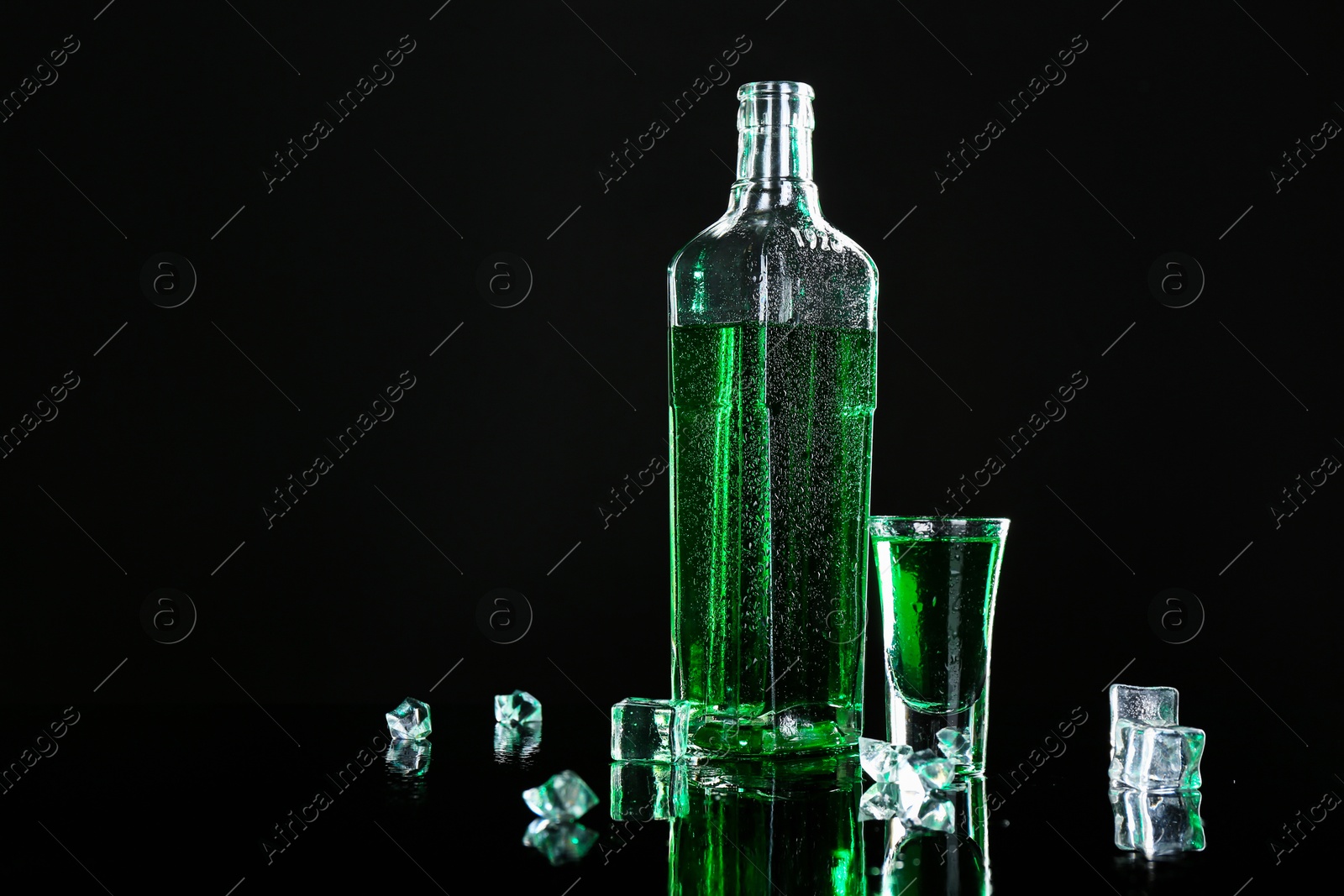 Photo of Absinthe in shot glass, bottle and ice cubes on black mirror surface, space for text