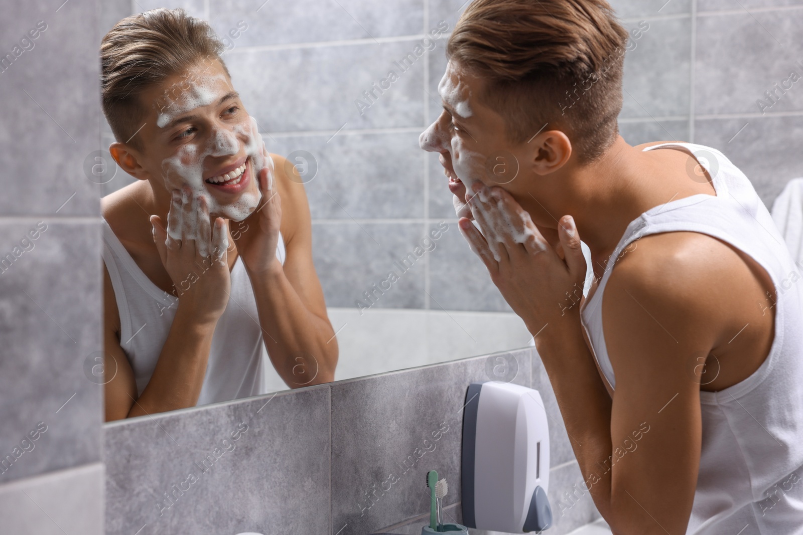 Photo of Man washing his face with cosmetic product near mirror in bathroom