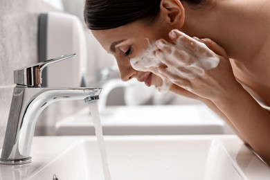 Photo of Woman washing her face with cleansing foam over sink in bathroom