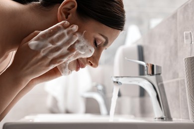 Photo of Woman washing her face with cleansing foam over sink in bathroom