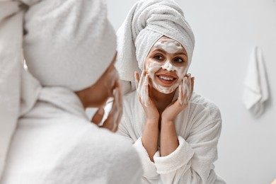 Photo of Woman washing her face with cleansing foam near mirror in bathroom