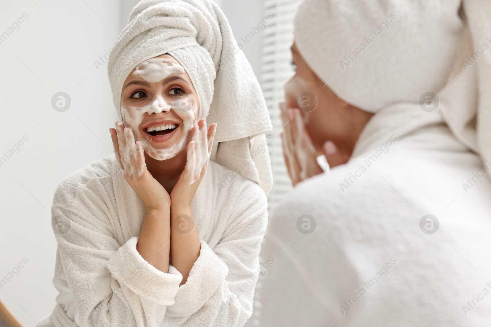 Photo of Woman washing her face with cleansing foam near mirror in bathroom