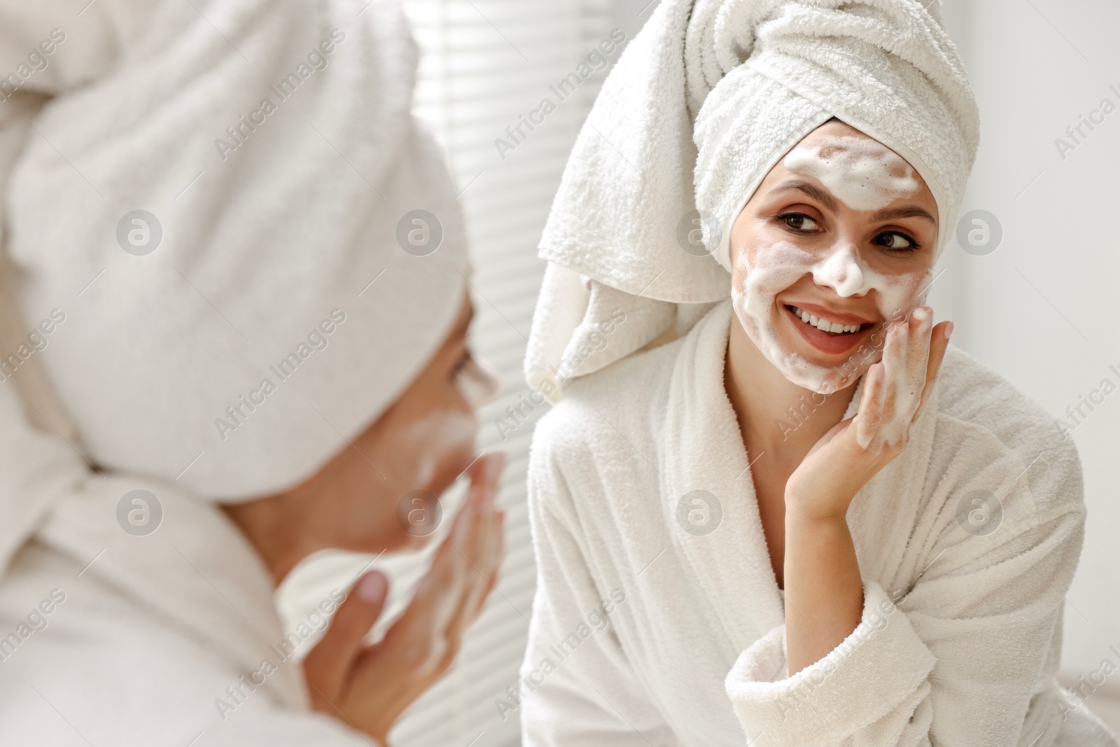 Photo of Woman washing her face with cleansing foam near mirror in bathroom