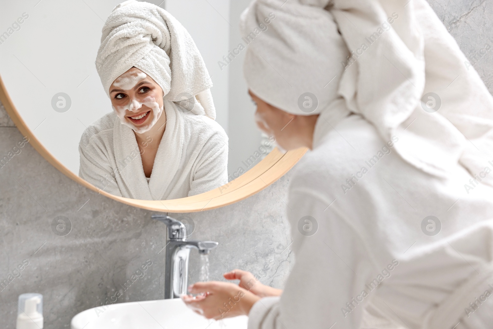 Photo of Woman washing her face with cleansing foam near mirror in bathroom