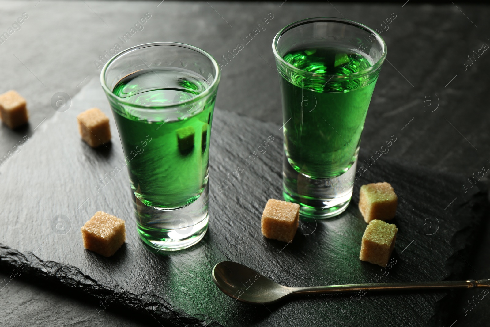 Photo of Delicious absinthe in shot glasses, spoon and brown sugar cubes on dark textured table, closeup