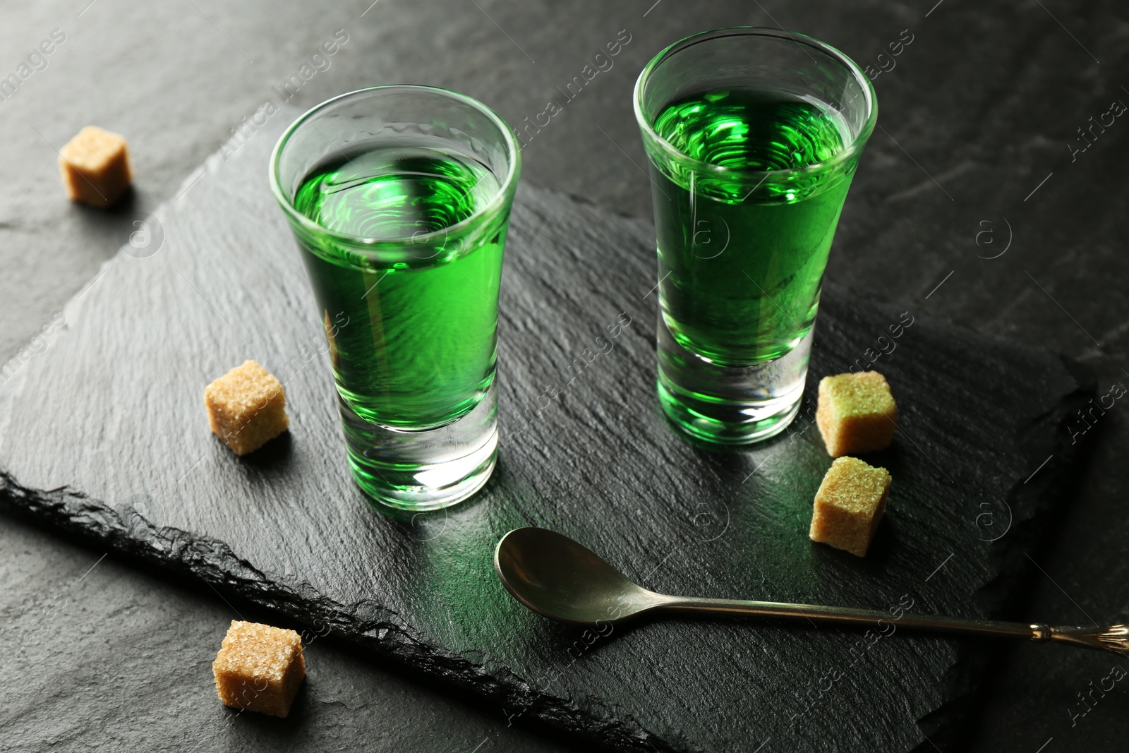 Photo of Delicious absinthe in shot glasses, spoon and brown sugar cubes on dark textured table, closeup