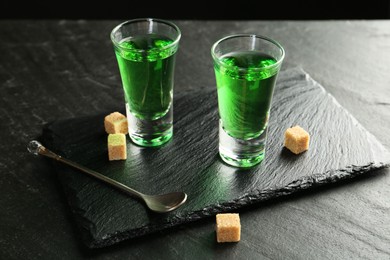 Photo of Delicious absinthe in shot glasses, spoon and brown sugar cubes on dark textured table, closeup