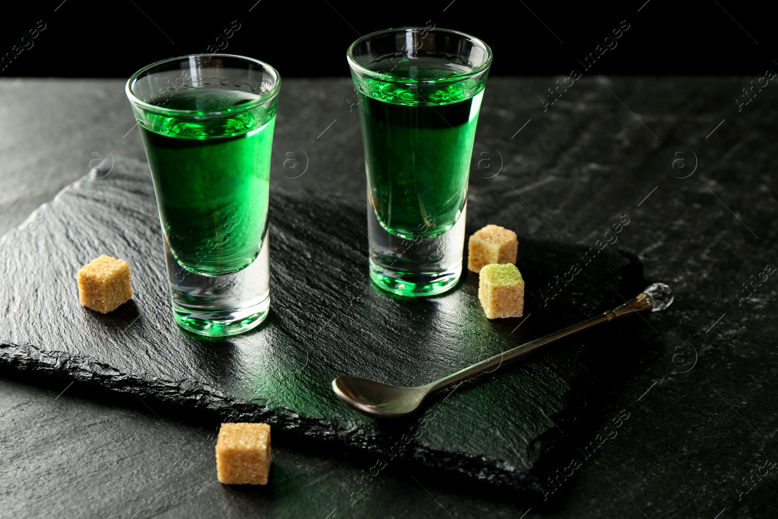 Photo of Delicious absinthe in shot glasses, spoon and brown sugar cubes on dark textured table, closeup