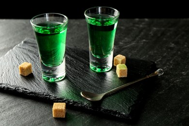 Photo of Delicious absinthe in shot glasses, spoon and brown sugar cubes on dark textured table, closeup