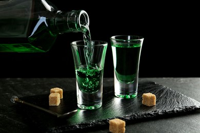 Photo of Pouring delicious absinthe from bottle into shot glass at grey textured table against black background, closeup