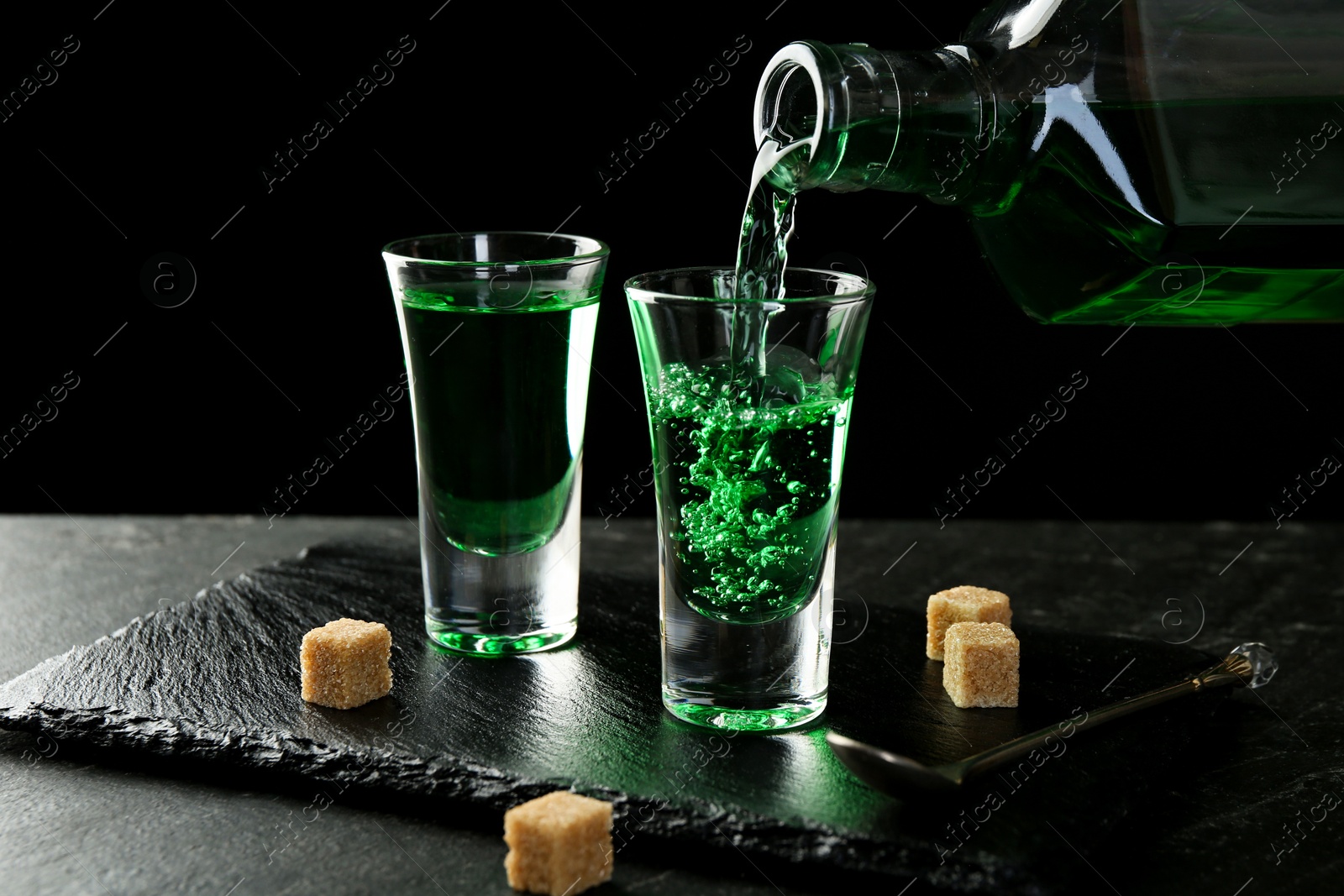 Photo of Pouring delicious absinthe from bottle into shot glass at grey textured table against black background, closeup