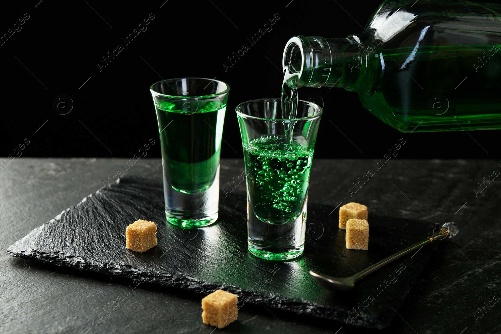 Photo of Pouring delicious absinthe from bottle into shot glass at grey textured table against black background, closeup