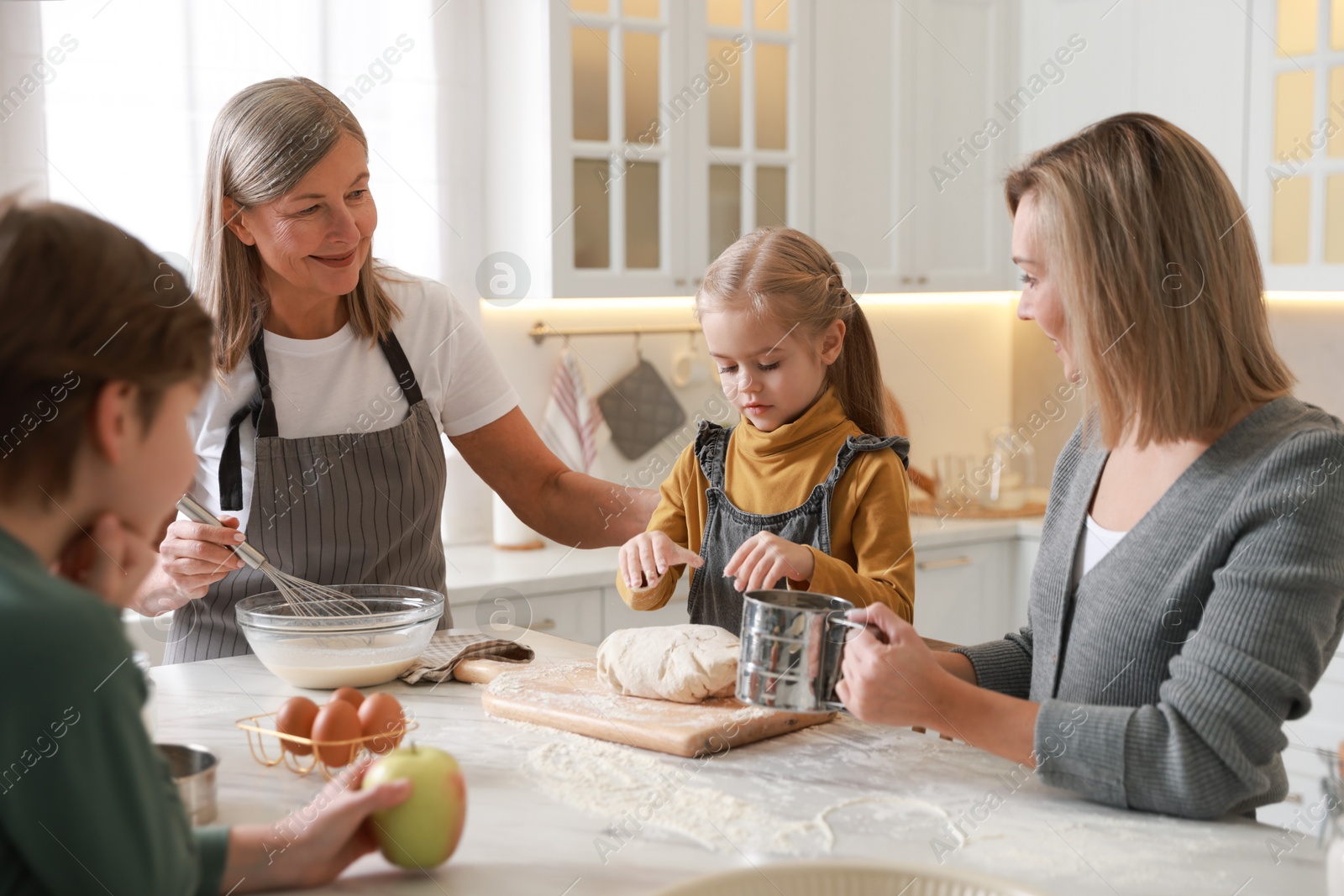 Photo of Little kids with their mother and grandmother making dough at white marble table in kitchen