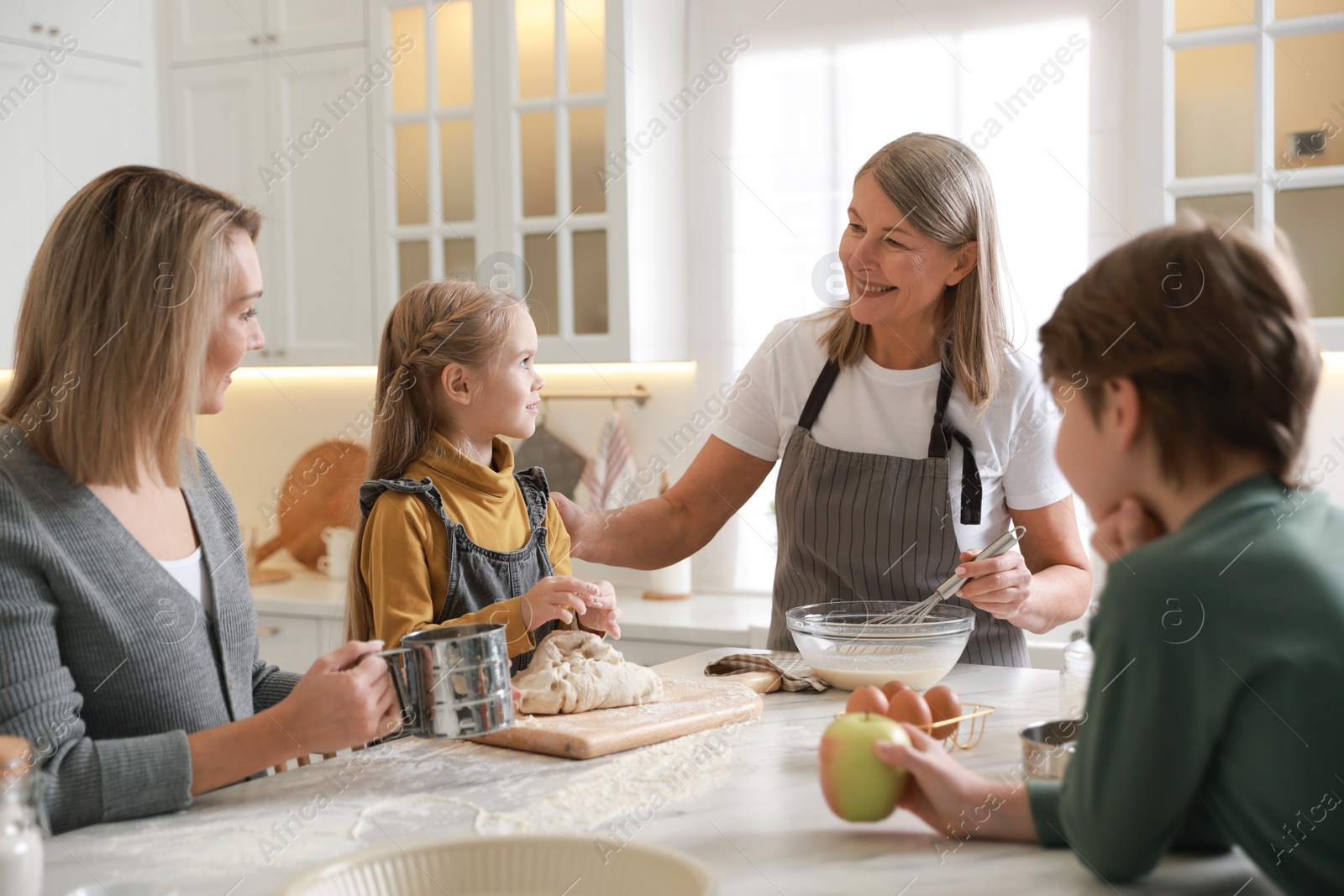 Photo of Little kids with their mother and grandmother making dough at white marble table in kitchen