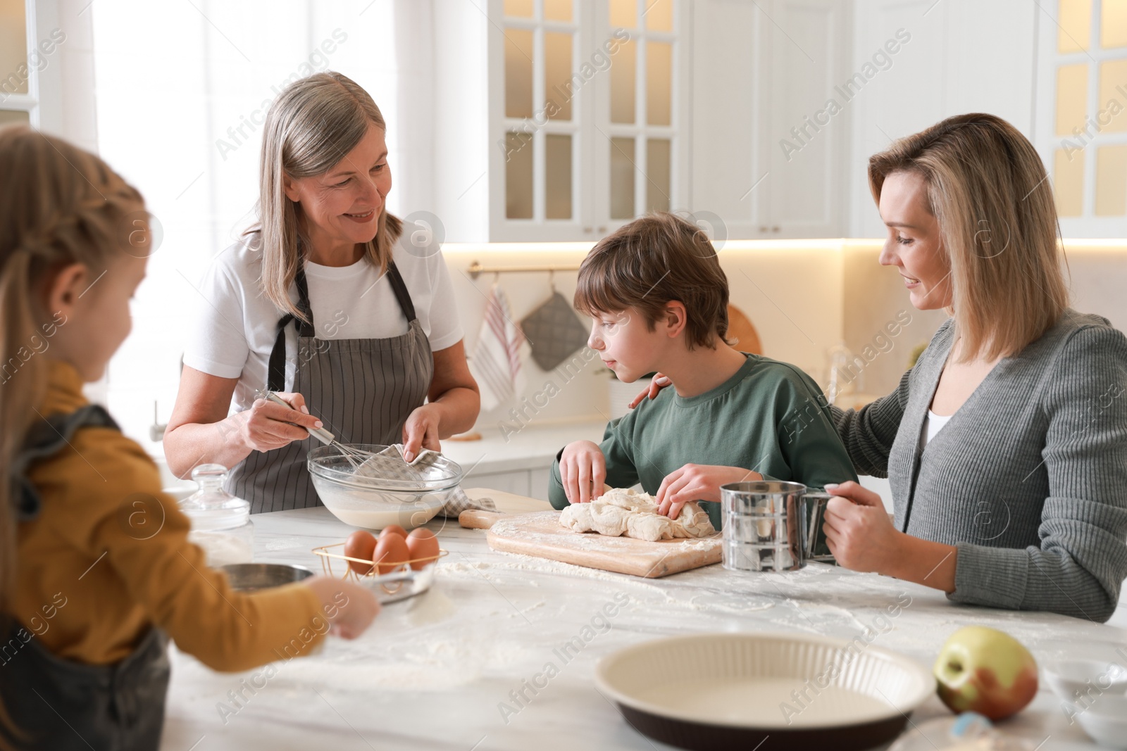 Photo of Little kids with their mother and grandmother making dough at white marble table in kitchen