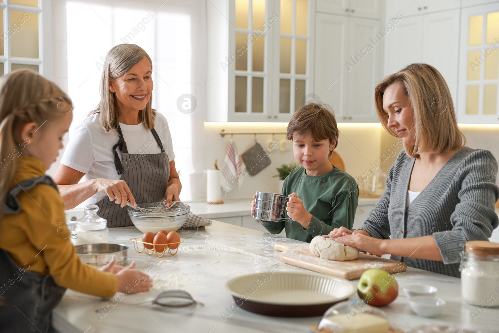 Photo of Little kids with their mother and grandmother making dough at white marble table in kitchen