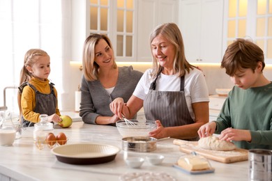 Photo of Little kids with their mother and grandmother making dough at white marble table in kitchen