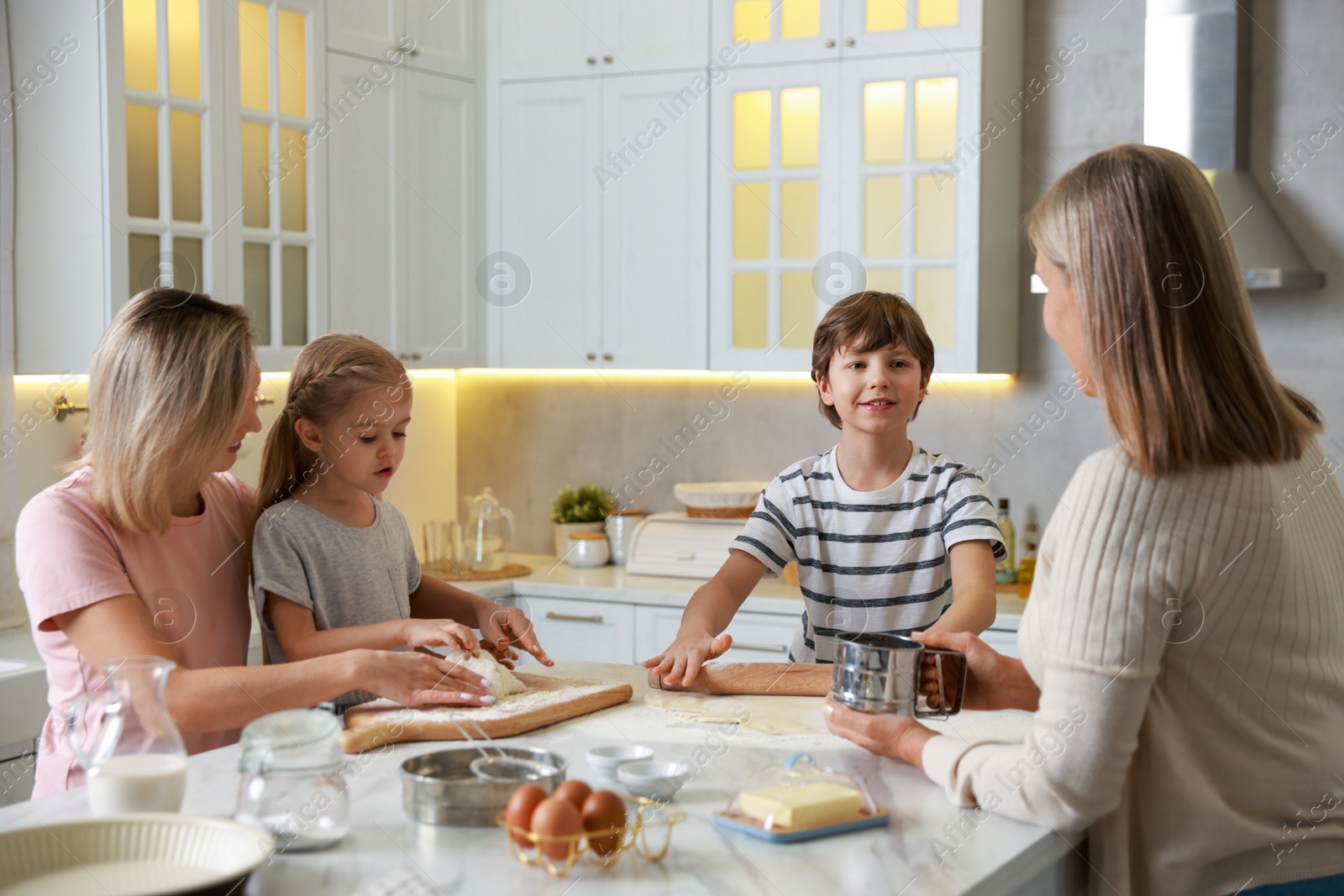 Photo of Little kids with their mother and grandmother making dough at white marble table in kitchen