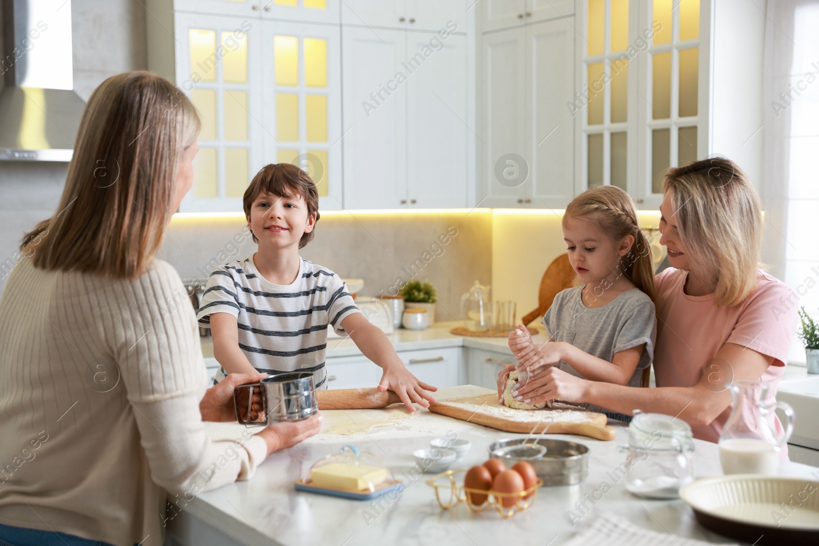 Photo of Little kids with their mother and grandmother making dough at white marble table in kitchen