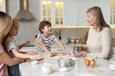 Photo of Little kids with their mother and grandmother making dough at white marble table in kitchen