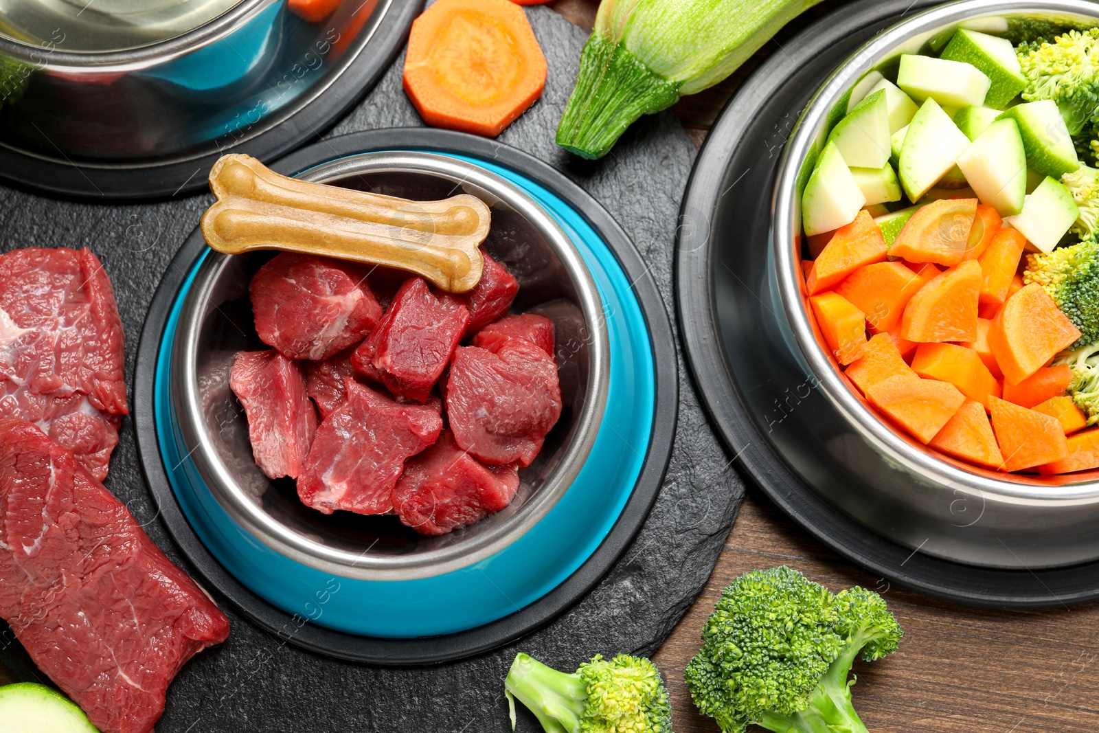 Photo of Pet food. Fresh beef in feeding bowl and chew bone among products on wooden table, flat lay