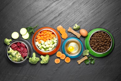 Photo of Natural and dry pet food in feeding bowls among products on dark textured table, flat lay