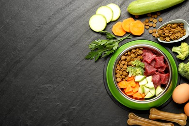 Photo of Natural and dry pet food in feeding bowl among products on dark textured table, flat lay. Space for text