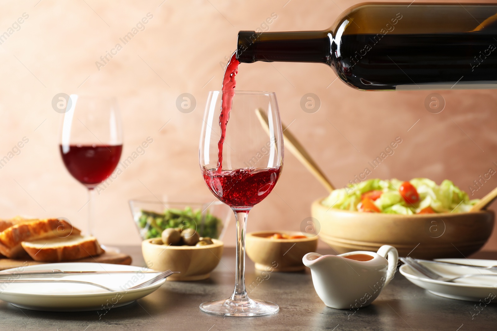 Photo of Pouring red wine into glass at grey table with tasty food, closeup