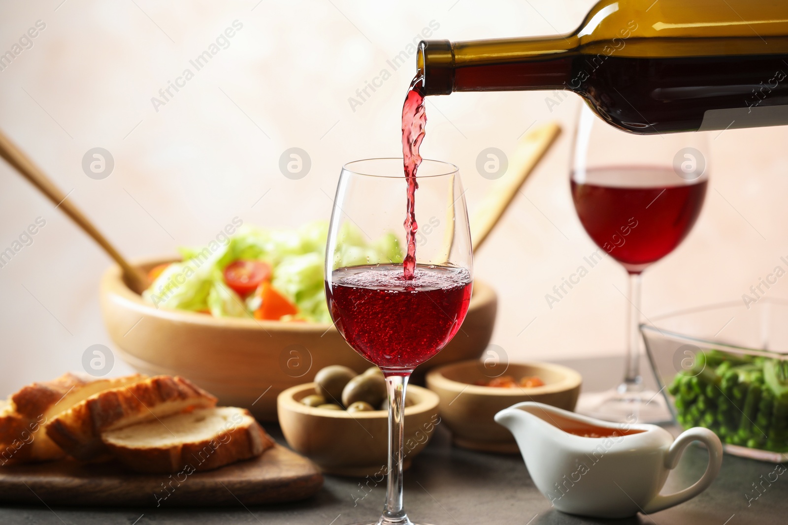 Photo of Pouring red wine into glass at grey table with tasty food, closeup