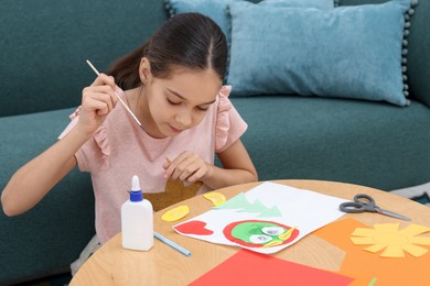 Photo of Girl applying glue onto paper figure for her creative project at table indoors. Art and craft