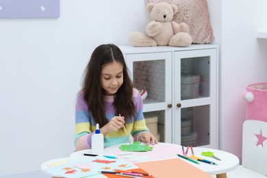 Photo of Girl applying glue onto paper figure for her creative project at table indoors. Art and craft