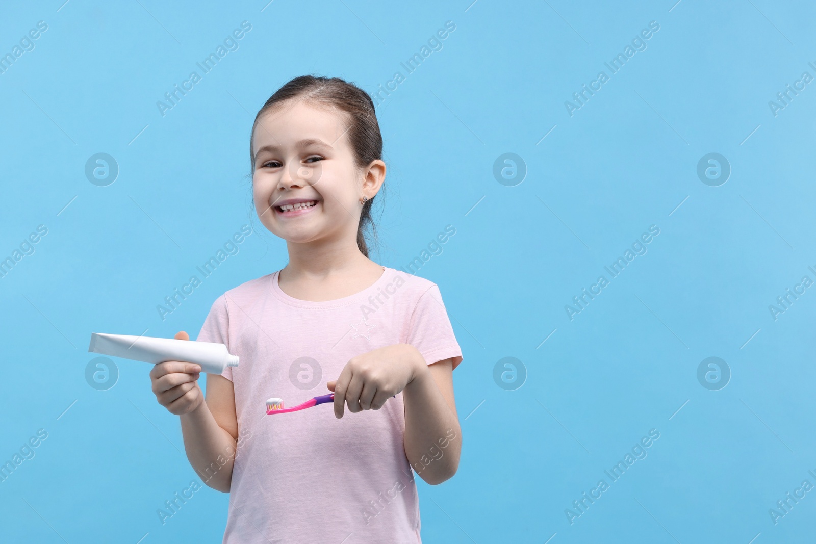 Photo of Cute girl with toothbrush and tube of toothpaste on light blue background, space for text