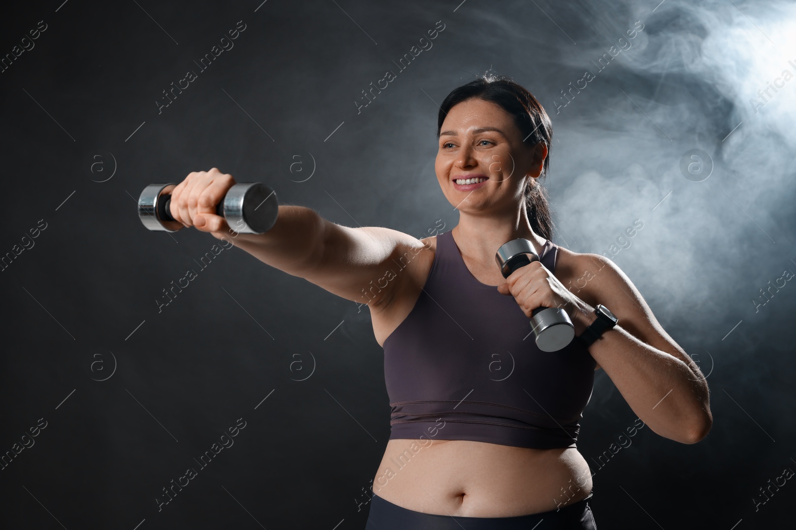 Photo of Plus size woman in gym clothes holding dumbbells on dark background with smoke