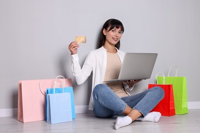 Photo of Internet shopping. Happy woman with credit card, laptop and colorful bags sitting near grey wall