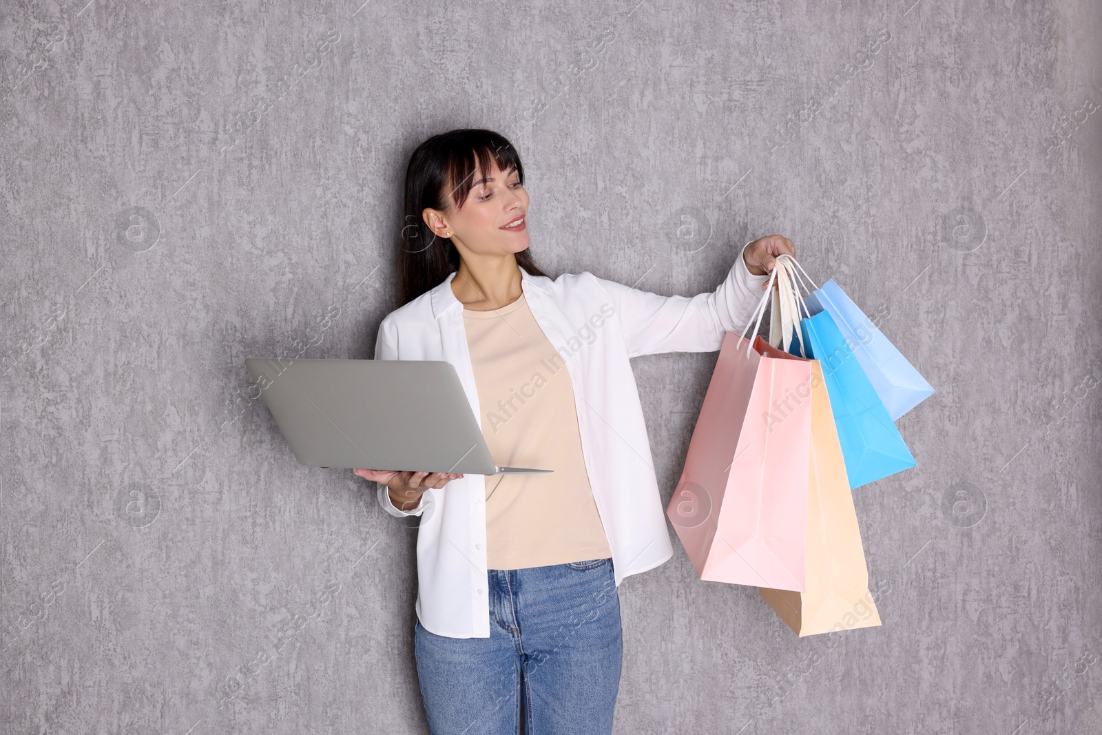 Photo of Internet shopping. Happy woman with laptop and colorful bags near grey wall