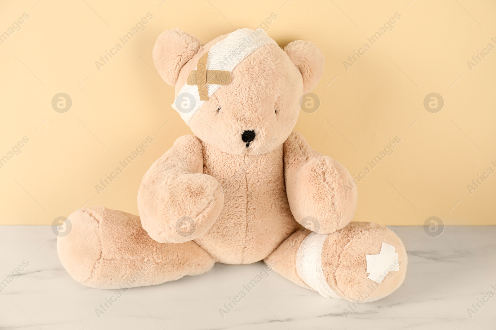 Teddy bear with bandages and sticking plasters on white marble table against beige background Photo of Teddy bear with bandages and sticking plasters on white marble table against beige background